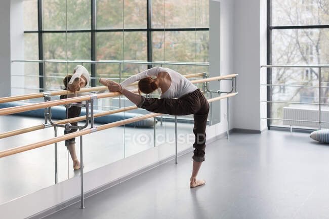 Young woman, ballerina doing stretching exercise near barre in rehearsal choreography studio — Stock Photo