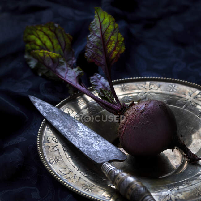 Целая свежая свекла с листьями помещена на винтажную пластину с кухонным ножом на черном фоне — стоковое фото