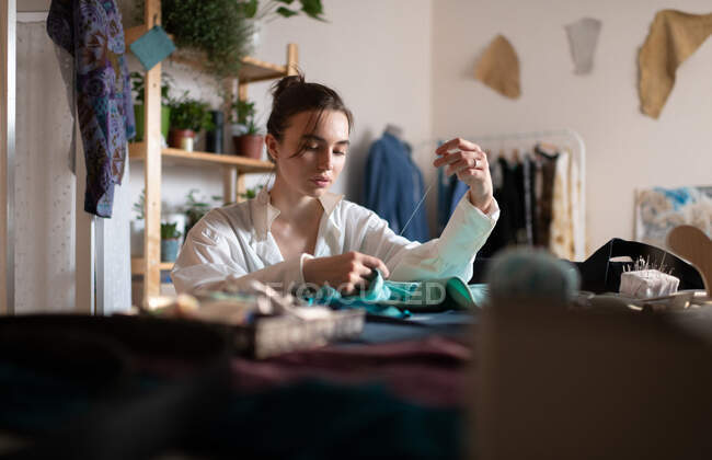 Young casual lady working on small business of dressmaking and sewing fabric pieces with needle in home studio — Stock Photo
