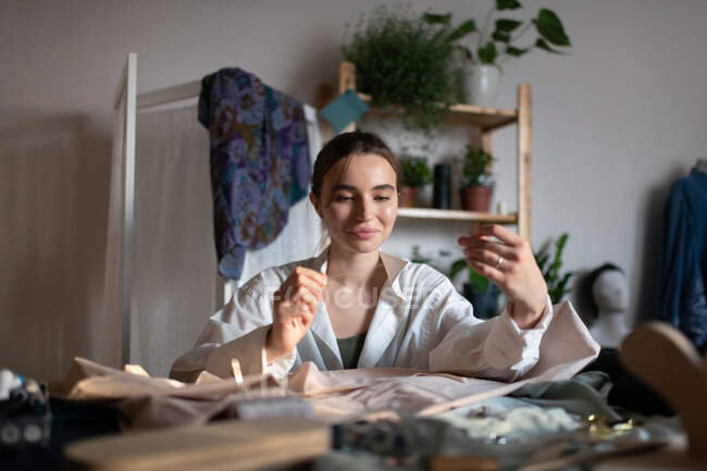 Encantador sastre femenino moderno sentado en la mesa en el estudio en casa que posee una pequeña empresa y coser ropa nueva - foto de stock