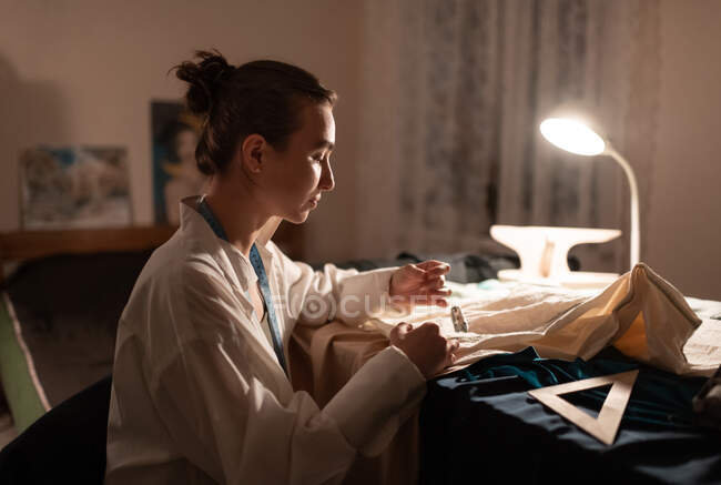 Vista laterale del sarto femminile calmo seduto a tavola con lampada accesa e pezzi di tessuto per cucire — Foto stock