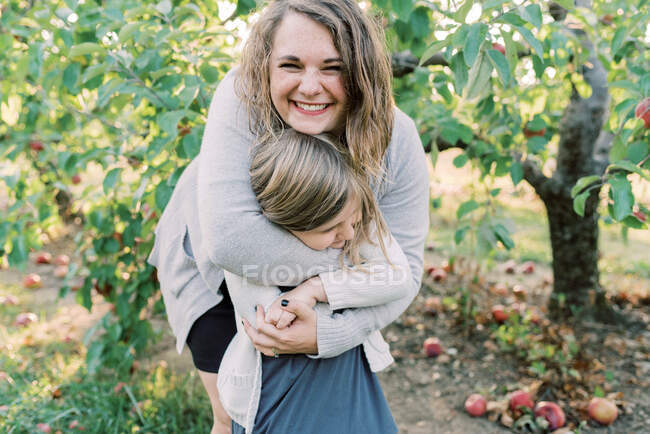 Uma mãe dando à filha um abraço de urso com árvores de maçãs nas costas — Fotografia de Stock