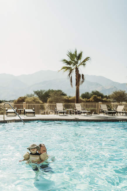Père et tout-petit fils nageant dans la piscine avec des montagnes et des palmiers — Photo de stock