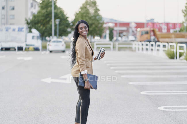 Retrato de una mujer negra con un traje marrón sosteniendo un portátil y un contenedor con café caminando por la calle - foto de stock