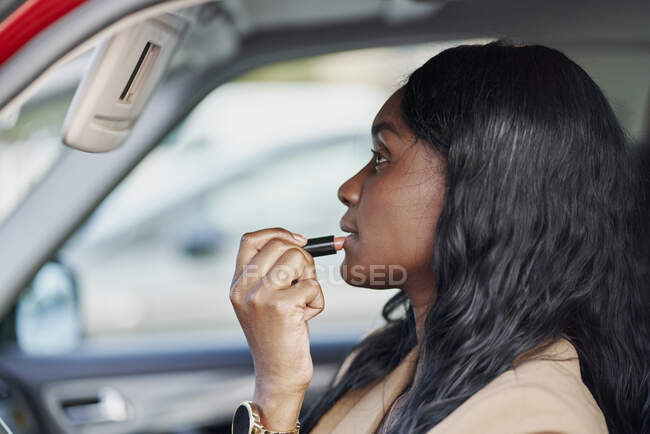 Portrait d'une femme noire en costume marron peignant ses lèvres à l'intérieur de sa voiture. Concept d'entreprise et élégance — Photo de stock