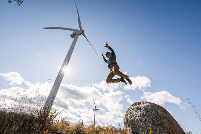 Человек прыгает из валуна с ветряной турбиной возвышающейся на заднем плане — стоковое фото