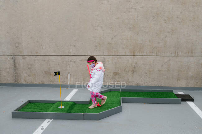 Menina em traje médico rosa e máscara facial jogando mini golfe — Fotografia de Stock