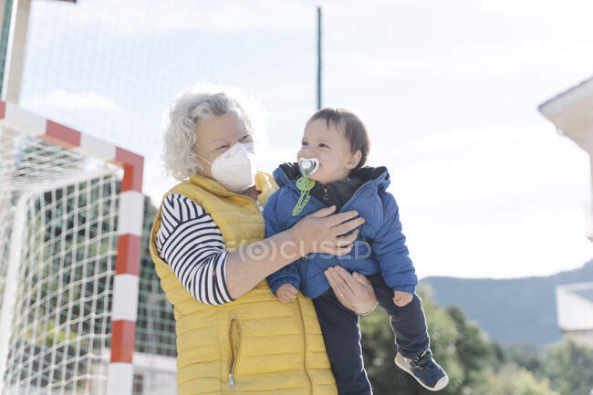 Abuela vistiendo una máscara médica con su nieto en sus brazos en un parque en un día soleado - foto de stock