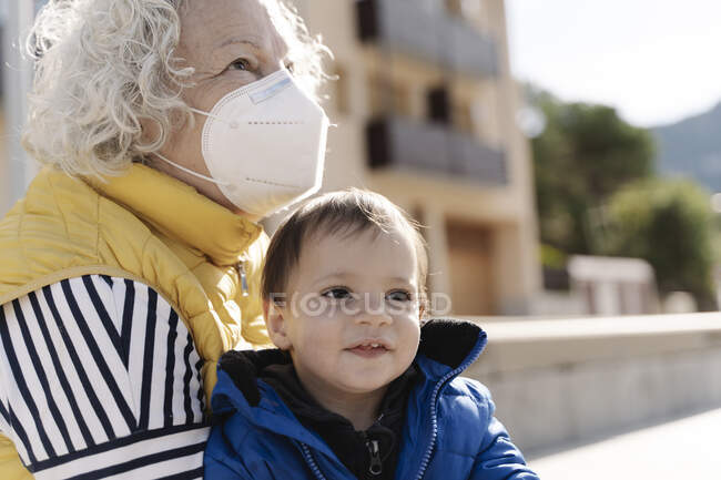 Retrato de la abuela con una máscara médica abrazando a su nieto en un parque - foto de stock