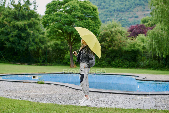 Joven mujer trenzada rubia con chaqueta de jean sentada en una piscina vacía con un paraguas. Concepto de moda - foto de stock