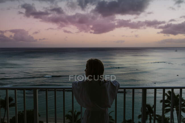 Jeune fille en peignoir regarde l'océan au coucher du soleil depuis le balcon à Hawaï — Photo de stock