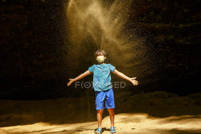 Um menino usando máscara facial fica em um desfiladeiro iluminado pelo sol jogando areia de ouro — Fotografia de Stock