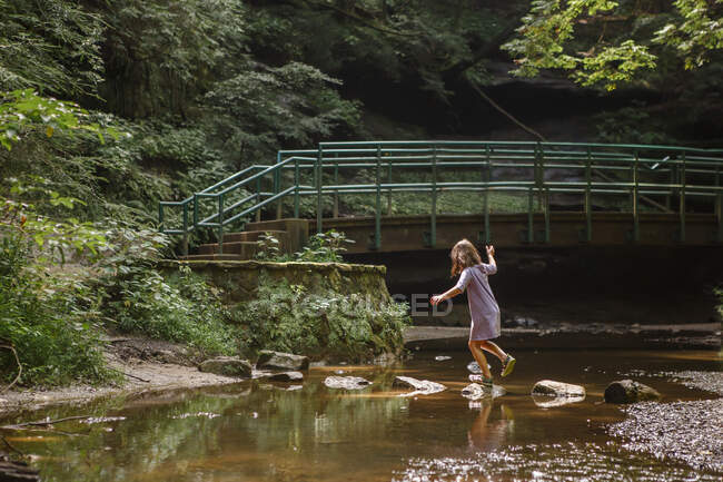 Маленька дівчинка балансує на ступінчастому камені через струмок у лісі — стокове фото