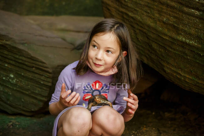 A smiling little girl crouches on ground with a painted turtle in lap — Stock Photo