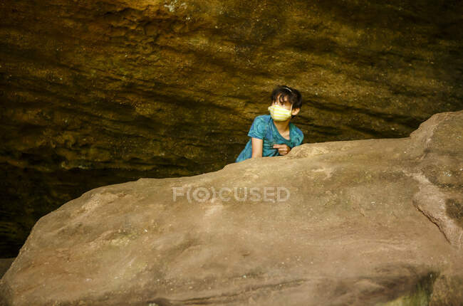 A boy wearing face masks peers over a large rock in a sandstone gorge — Stock Photo