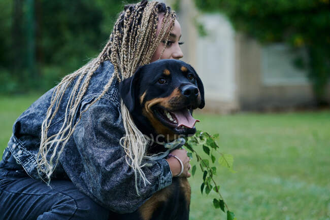 Young woman with blonde colored braided hair wearing a denim jacket and pants hugging her dog — Stock Photo