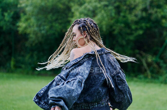 Una giovane donna con i capelli biondi intrecciati che indossa una giacca e jeans muove le trecce di fronte alla fotocamera — Foto stock
