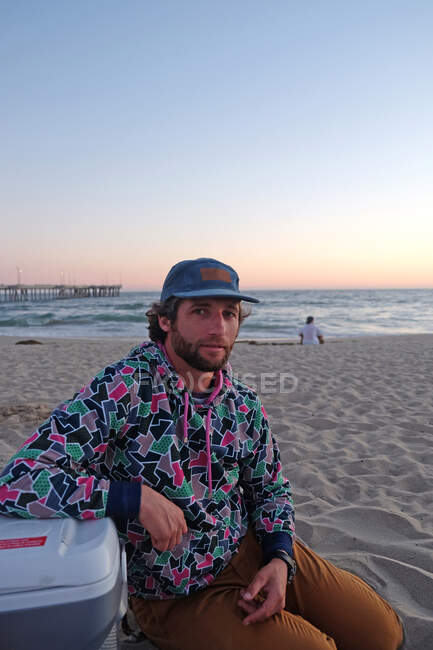 Homem vestindo um boné com barba e cabelo encaracolado sentado na praia ao pôr do sol — Fotografia de Stock