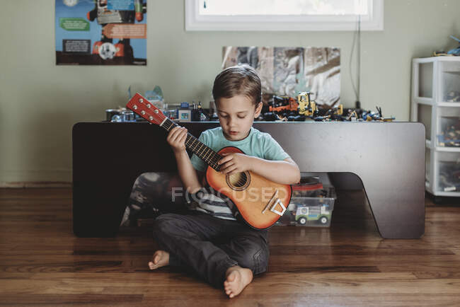 Niño de 5 años sentado en el suelo de madera tocando la guitarra - foto de stock