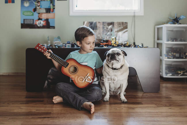 Barefoot boy holding guitar sitting next to pet Pug on hardwood floor — Stock Photo