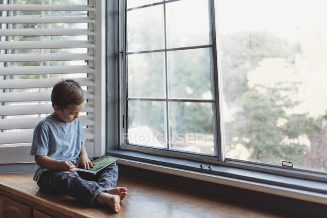 Junge liest in Fensternische mit weichem Licht und Rollläden — Stockfoto