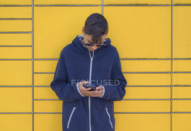Підліток з сонцезахисними окулярами використовує свій мобільний телефон. Жовта стіна — стокове фото