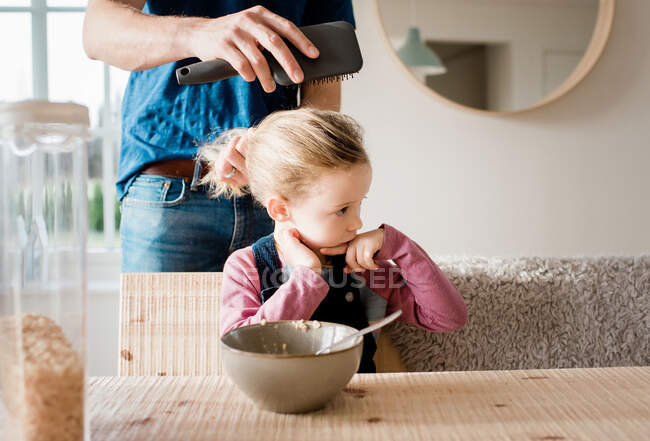 Father brushing his daughters hair at breakfast table before school — Stock Photo