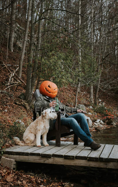 Uomo con la testa di zucca spaventosa per Halloween seduto sul molo con il cane. — Foto stock