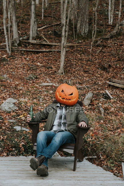 Uomo con la testa di zucca spaventosa ad Halloween seduto sul molo con la birra. — Foto stock