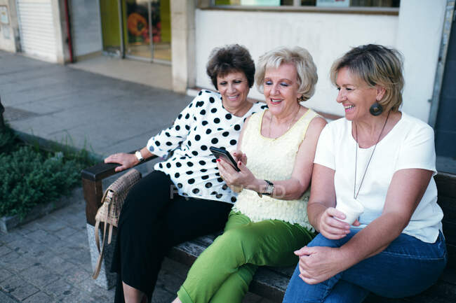 Зрелые женщины улыбаются на мобильный телефон, сидя на скамейке — стоковое фото