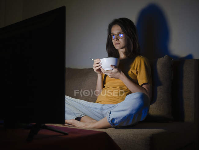 Jovem mulher assistindo tv com tigela de comida em seu quarto à noite — Fotografia de Stock