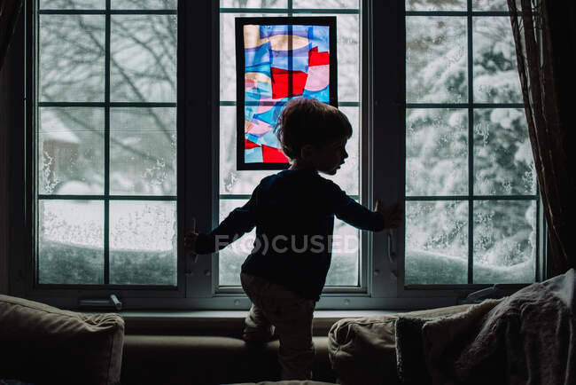Um menino fica em frente a uma janela nevada. — Fotografia de Stock