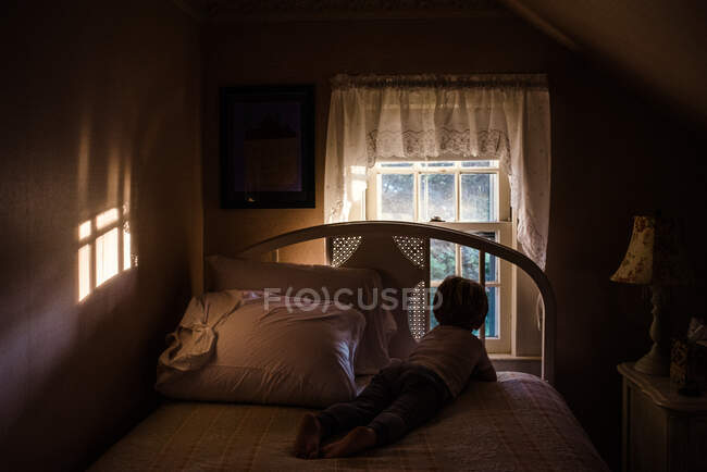 Un niño se acuesta en una cama y mira por una ventana. - foto de stock