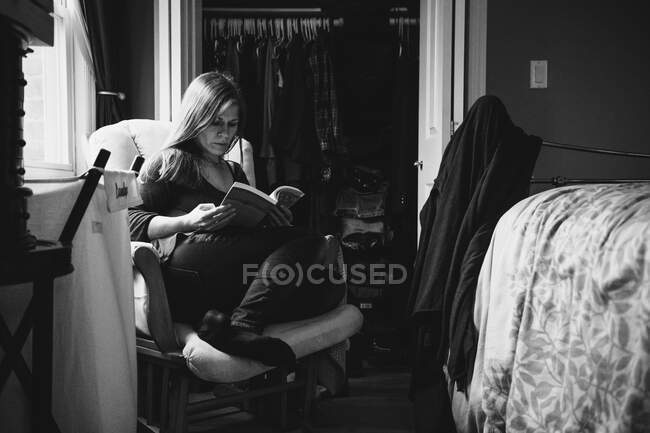 Una mujer se sienta en una silla leyendo un libro. - foto de stock