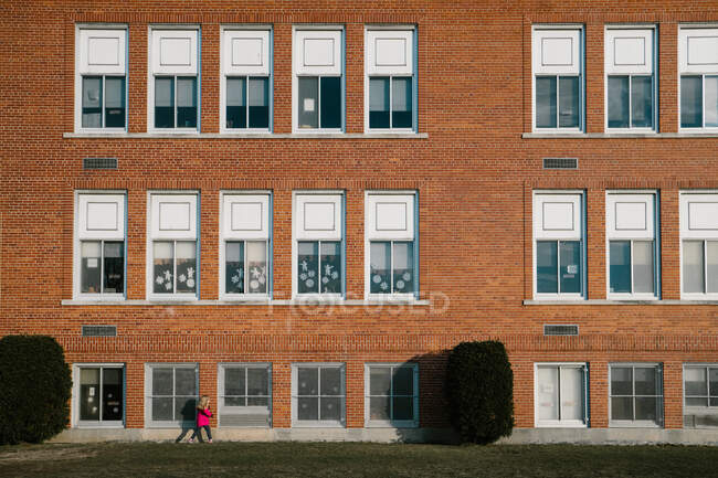 Маленькая девочка в розовом пальто проходит мимо школьного здания. — стоковое фото