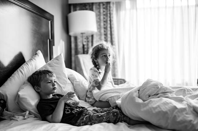 Two children lie in a hotel room bed eating snacks. — Stock Photo