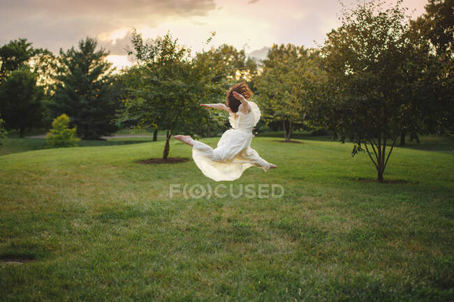 Dancer in long white dress twirling in golden light in park at sunset — Stock Photo