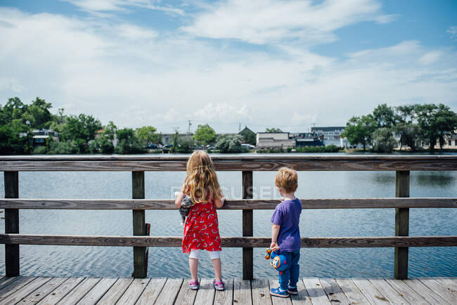 Two children stand at a pier and look out at a pond. — Stock Photo