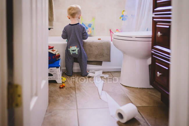 Um menino fica em uma casa de banho com um rolo de toalete desfraldado. — Fotografia de Stock