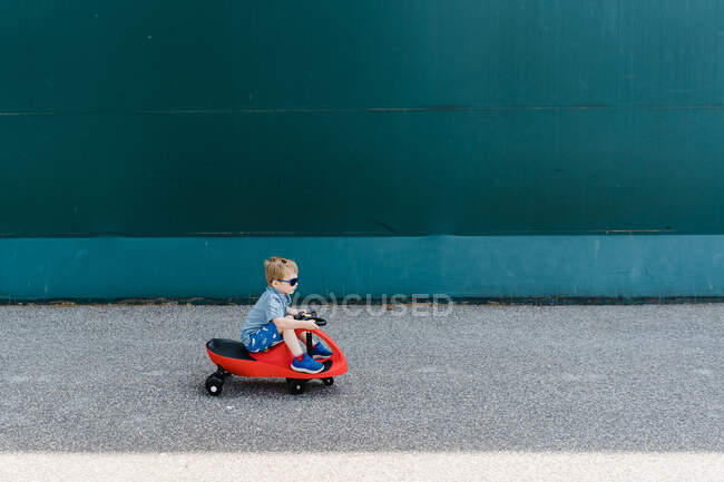 Um menino monta um carro de plasma vermelho na frente de uma parede verde. — Fotografia de Stock