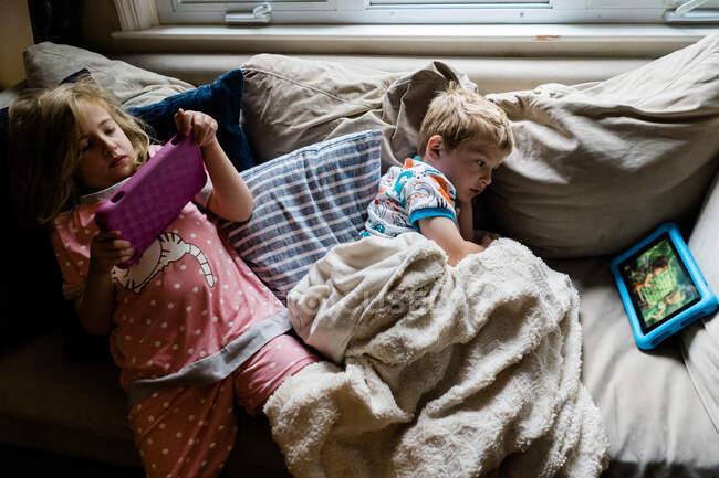 Двое детей лежат на диване и смотрят таблетки. — стоковое фото