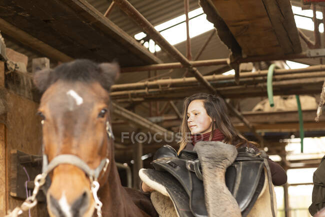 Jovem mulher com um cavalo em um estábulo de cavalo com uma sela — Fotografia de Stock