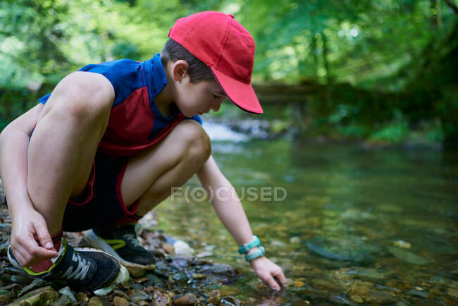 Bambino con cappello rosso sulla riva del fiume che gioca con acqua e pietre nella foresta — Foto stock