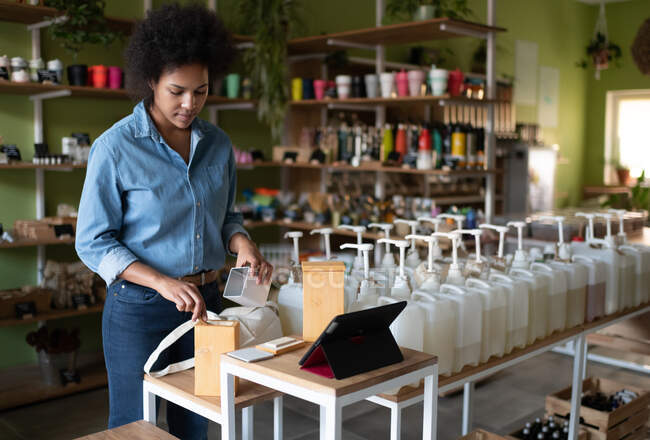 Змішана раса жінка заповнює багаторазову коробку з продуктом для онлайн-замовлення в еко-магазині — стокове фото