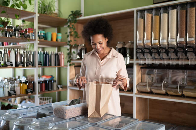 Delighted ethic woman looking inside paper bag during work in eco grocery shop — Stock Photo