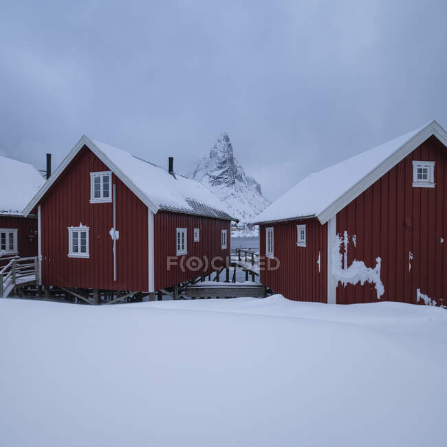 Горная вершина Ольстинд и каюты красного цвета, возвышающиеся над заснеженным побережьем, Лофские острова, Норвегия — стоковое фото