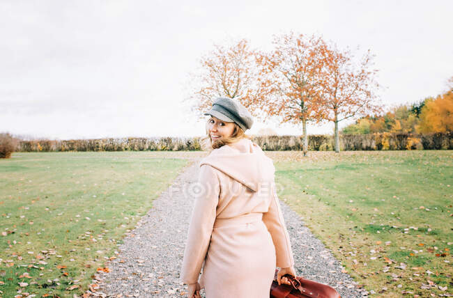 Frau geht eine Landstraße entlang und blickt lächelnd mit einem Koffer zurück — Stockfoto