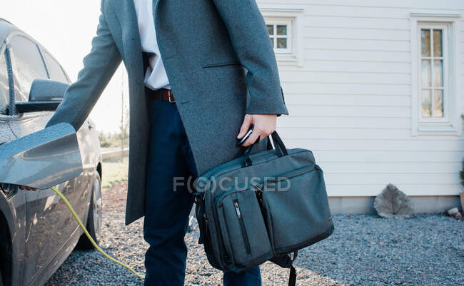 Uomo scollegare auto elettrica tenendo la borsa da lavoro lasciando casa — Foto stock