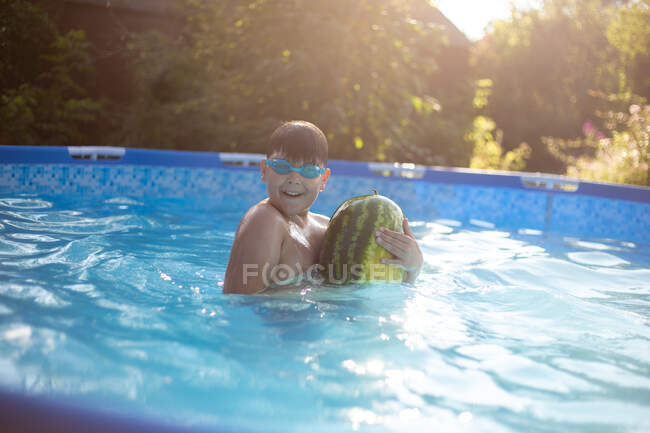 Chico divertido nada y juega con sandía en la piscina - foto de stock