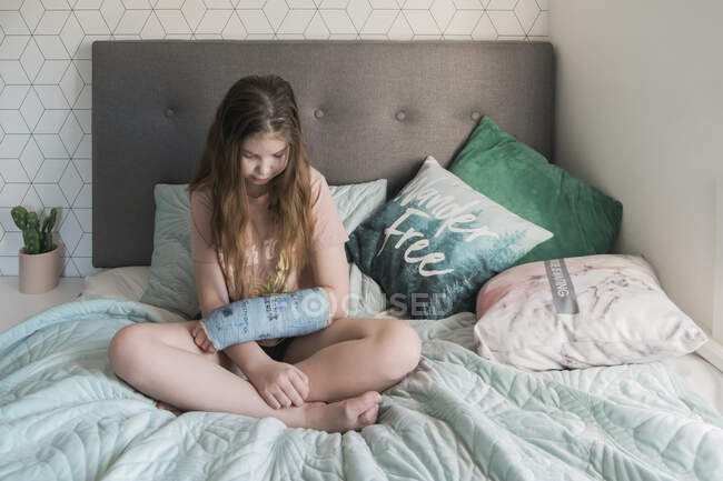Молода дівчина сидить на ліжку, дивлячись на її руку в синій гіпсовий литий — стокове фото